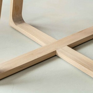 May include: Close-up of the base of a wooden table with a cross-shaped design. The table legs are made of light-colored wood and have a smooth finish.
