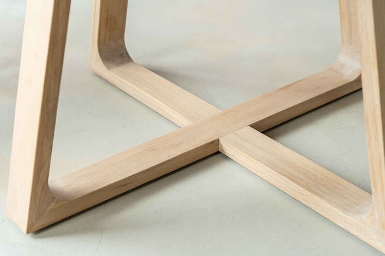 May include: Close-up of the base of a wooden table with a cross-shaped design. The table legs are made of light-colored wood and have a smooth finish.