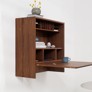 Peut inclure: Un bureau mural en bois avec une surface de bureau rabattable. Le bureau comporte plusieurs étagères et compartiments de rangement. Une tasse bleue est posée sur la surface du bureau. Un petit vase avec des fleurs jaunes est posé dessus.