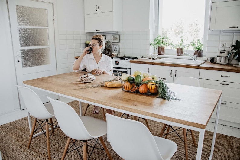 May include: A bright, airy kitchen with a wooden table and white chairs. A wire basket holds fresh produce. A person is seated at the table, enjoying a drink. The room features white cabinets, a window, and a rug.
