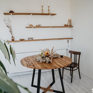 May include: A round wooden dining table with a black metal base, surrounded by a white wall with wooden shelves. The table is decorated with a bouquet of dried flowers and has a natural wood finish. The table seats four people. "Seats 4" is written on the floor.