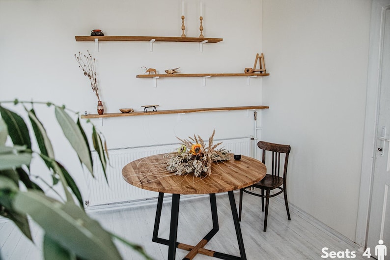 May include: A round wooden dining table with a black metal base, surrounded by a white wall with wooden shelves. The table is decorated with a bouquet of dried flowers and has a natural wood finish. The table seats four people. "Seats 4" is written on the floor.
