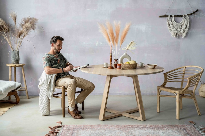May include: A round wooden table with a natural finish, surrounded by three chairs. One chair is made of woven rattan, and the other two are wooden. A man is sitting at the table, writing in a notebook. The table is decorated with dried pampas grass, candles, and a bowl of fruit.