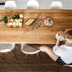 May include: A wooden dining table with a rectangular shape, set with a wire basket of fruit, corn, and bread. White chairs surround the table, and a person is seated at the table. The table is set on a brown woven rug.