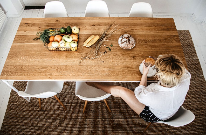 May include: A wooden dining table with a rectangular shape, set with a wire basket of fruit, corn, and bread. White chairs surround the table, and a person is seated at the table. The table is set on a brown woven rug.