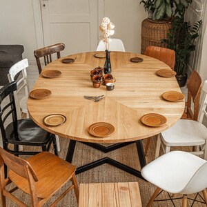 May include: A round wooden dining table with a black metal base, surrounded by chairs in various styles and colors. The table is set with plates and cups, and a vase of dried flowers sits in the center.