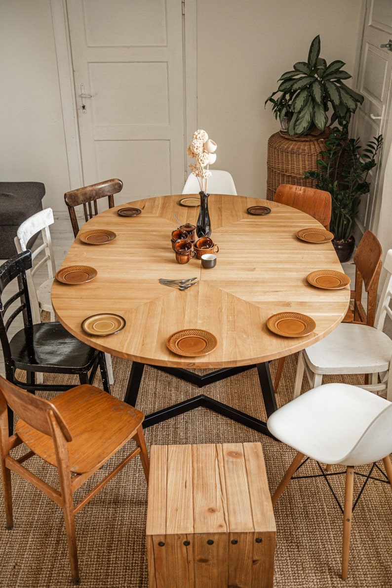 May include: A round wooden dining table with a black metal base, surrounded by chairs in various styles and colors. The table is set with plates and cups, and a vase of dried flowers sits in the center.