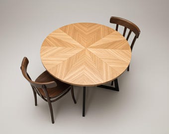 May include: A round wooden table with a light brown, chevron-patterned top. Two dark brown wooden chairs with curved backs are positioned around the table. The table has black metal legs.