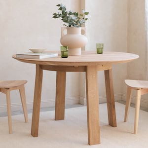 May include: A round wooden dining table with four legs and two matching chairs. The table has a light wood finish and the chairs have a natural wood finish. A vase with green leaves and a light brown ceramic vase is on the table.