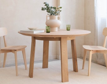 May include: A round wooden dining table with four legs and two matching chairs. The table has a light wood finish and the chairs have a natural wood finish. A vase with green leaves and a light brown ceramic vase is on the table.