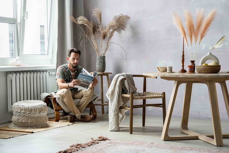 May include: A man sits in a chair reading a magazine in a living room with a wooden table, two chairs, and a vase of dried pampas grass.