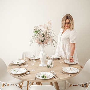May include: A round wooden table with white chairs set for a dinner party. The table is decorated with a white vase filled with dried flowers and greenery.