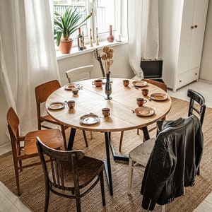 May include: A wooden dining table with six chairs, set with plates, cups, and saucers. A vase with dried flowers sits in the center of the table. A leather jacket hangs on the back of a chair.