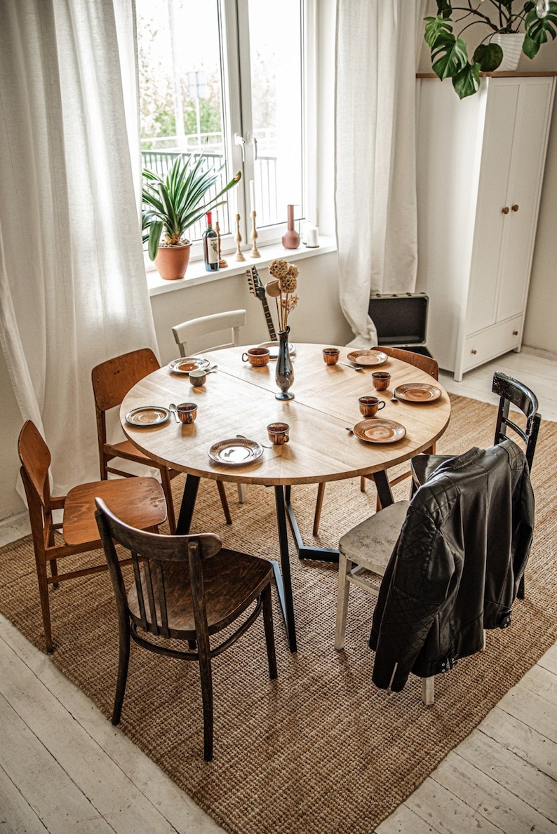 May include: A wooden dining table with six chairs, set with plates, cups, and saucers. A vase with dried flowers sits in the center of the table. A leather jacket hangs on the back of a chair.