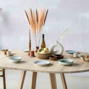 May include: A round wooden dining table with a light wood finish. The table is set with plates, bowls, and glasses. There are also some decorative items on the table, including a vase with dried flowers, a bottle of wine, and a bowl of fruit. The table is surrounded by chairs with woven seats.