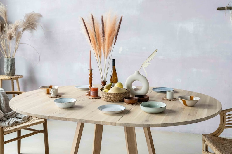 May include: A round wooden dining table with a light wood finish. The table is set with plates, bowls, and glasses. There are also some decorative items on the table, including a vase with dried flowers, a bottle of wine, and a bowl of fruit. The table is surrounded by chairs with woven seats.