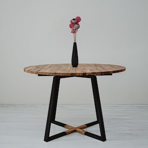 May include: A round wooden table with a black metal base. The table has a natural wood finish and the base is made of a sturdy, industrial-style metal. A vase with pink and gray flowers sits on the table.