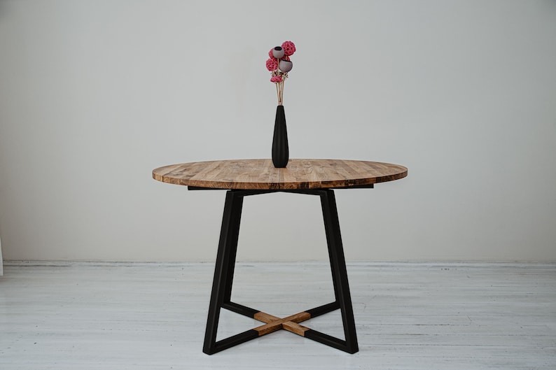 May include: A round wooden table with a black metal base. The table has a natural wood finish and the base is made of a sturdy, industrial-style metal. A vase with pink and gray flowers sits on the table.