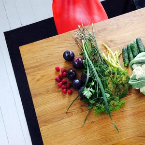 May include: Overhead shot of a wooden table with fresh produce. The arrangement includes dark purple plums, bright red raspberries, green beans, cucumbers, cauliflower, and herbs. A red chair is visible in the background.