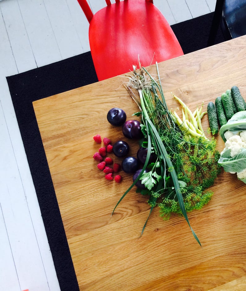 May include: Overhead shot of a wooden table with fresh produce. The arrangement includes dark purple plums, bright red raspberries, green beans, cucumbers, cauliflower, and herbs. A red chair is visible in the background.