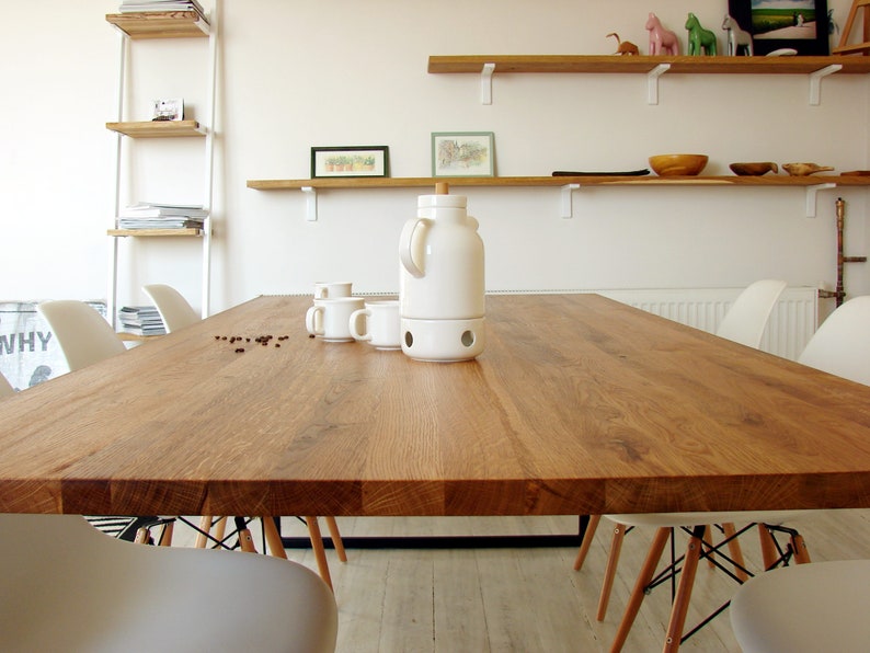 May include: A large, rectangular wooden dining table with a natural finish. The table is surrounded by white chairs with wooden legs and black metal supports. A white ceramic coffee pot and cups sit on the table, along with scattered coffee beans. Shelves with decor are visible in the background.