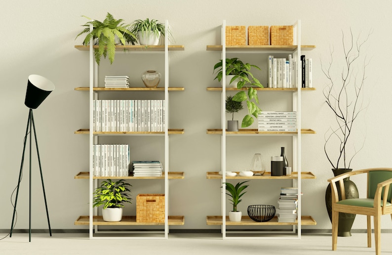Puede incluir: Dos estanter&iacute;as de metal blanco con estantes de madera, llenas de libros, plantas y objetos decorativos. Las estanter&iacute;as est&aacute;n contra una pared blanca con una l&aacute;mpara de pie de tr&iacute;pode negra a la izquierda.