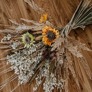 May include: A bouquet of dried flowers with sunflowers, pampas grass, and other dried grasses. The bouquet is arranged on a wooden surface.