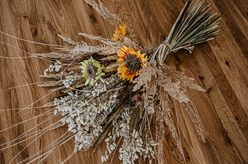 May include: A bouquet of dried flowers with sunflowers, pampas grass, and other dried grasses. The bouquet is arranged on a wooden surface.