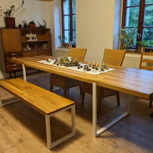 May include: A long wooden dining table with white metal legs, a matching wooden bench, and four chairs. The table is set with a white tablecloth, a centerpiece of greenery and candles, and a variety of decorative items.