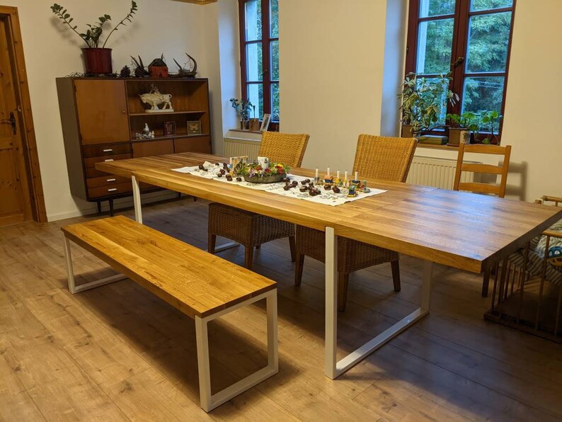 May include: A long wooden dining table with white metal legs, a matching wooden bench, and four chairs. The table is set with a white tablecloth, a centerpiece of greenery and candles, and a variety of decorative items.