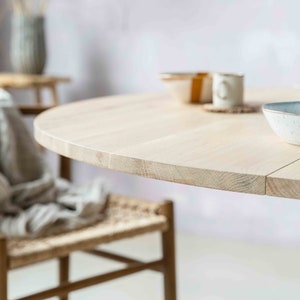 May include: A light wood round table with a natural finish. The table is set with a white bowl and a cup. The table is in a room with a light pink wall.