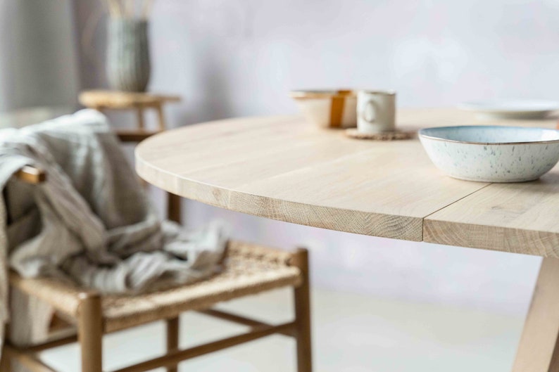 May include: A light wood round table with a natural finish. The table is set with a white bowl and a cup. The table is in a room with a light pink wall.