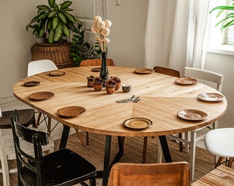 Könnte beinhalten: Ein runder Esstisch aus Holz mit schwarzen Metallbeinen, gedeckt mit Tellern und Besteck. Der Tisch ist von Stühlen umgeben, darunter ein schwarzer Stuhl mit einem geflochtenen Sitz und ein weißer Stuhl mit einem geflochtenen Sitz.