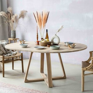 May include: A light wood oval dining table with a cross-shaped base. The table is set with plates, bowls, and a vase of dried pampas grass. Two chairs are visible, one with a woven rattan seat and the other with a woven fabric seat.