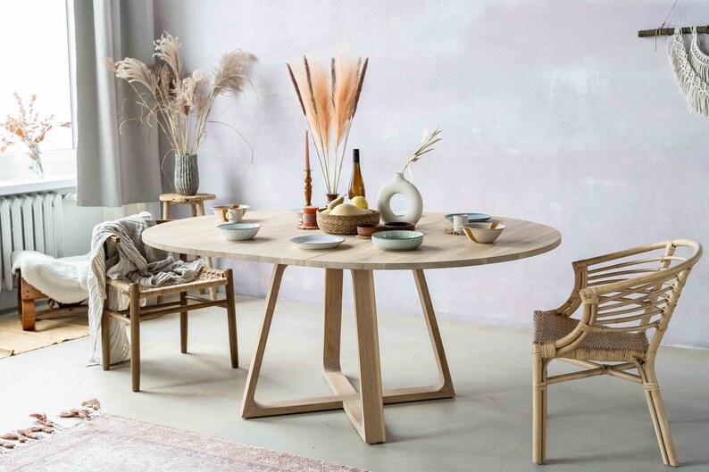May include: A light wood oval dining table with a cross-shaped base. The table is set with plates, bowls, and a vase of dried pampas grass. Two chairs are visible, one with a woven rattan seat and the other with a woven fabric seat.
