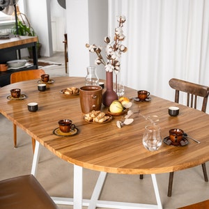 K&ouml;nnte beinhalten: Ein ovaler Holztisch mit wei&szlig;en Beinen, gedeckt f&uuml;r eine Mahlzeit mit braunen und wei&szlig;en Tellern, einem Glas Wasser und einer Vase mit wei&szlig;en Baumwollblumen.