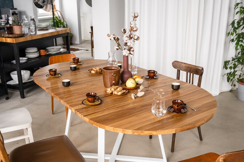 K&ouml;nnte beinhalten: Ein ovaler Holztisch mit wei&szlig;en Beinen, gedeckt f&uuml;r eine Mahlzeit mit braunen und wei&szlig;en Tellern, einem Glas Wasser und einer Vase mit wei&szlig;en Baumwollblumen.