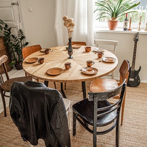 May include: A round wooden dining table with six chairs, set with plates, cups, and saucers. A vase with dried flowers sits in the center of the table. A guitar leans against the wall behind the table.