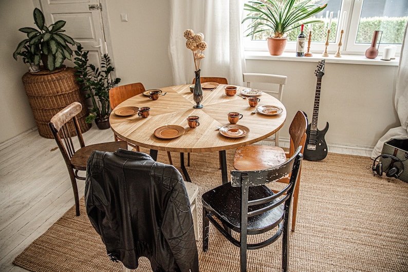 May include: A round wooden dining table with six chairs, set with plates, cups, and saucers. A vase with dried flowers sits in the center of the table. A guitar leans against the wall behind the table.
