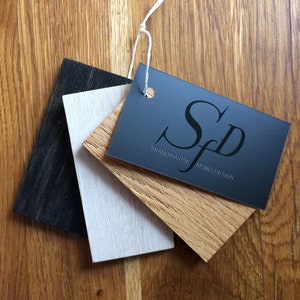 May include: A collection of wood samples in black, white, and natural wood tones, with a dark gray tag attached by a white string. The tag reads "Skandinavisk Mobeldesign" in white lettering. The samples are arranged on a wooden surface.