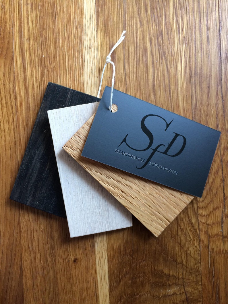 May include: A collection of wood samples in black, white, and natural wood tones, with a dark gray tag attached by a white string. The tag reads "Skandinavisk Mobeldesign" in white lettering. The samples are arranged on a wooden surface.