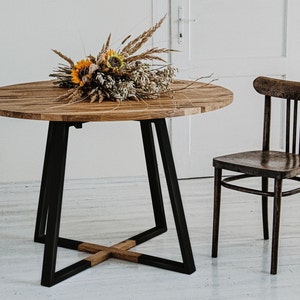 May include: A round wooden table with black metal legs and a cross-shaped support. The table is topped with a bouquet of dried flowers and is set against a white wall.