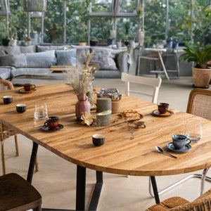 K&ouml;nnte beinhalten: Ein ovaler Holztisch mit schwarzen Metallbeinen, gedeckt mit vier St&uuml;hlen und einer Auswahl an Geschirr, darunter Tassen, Untertassen und ein Glas Wasser. Der Tisch ist von Gr&uuml;npflanzen und einer Couch im Hintergrund umgeben.