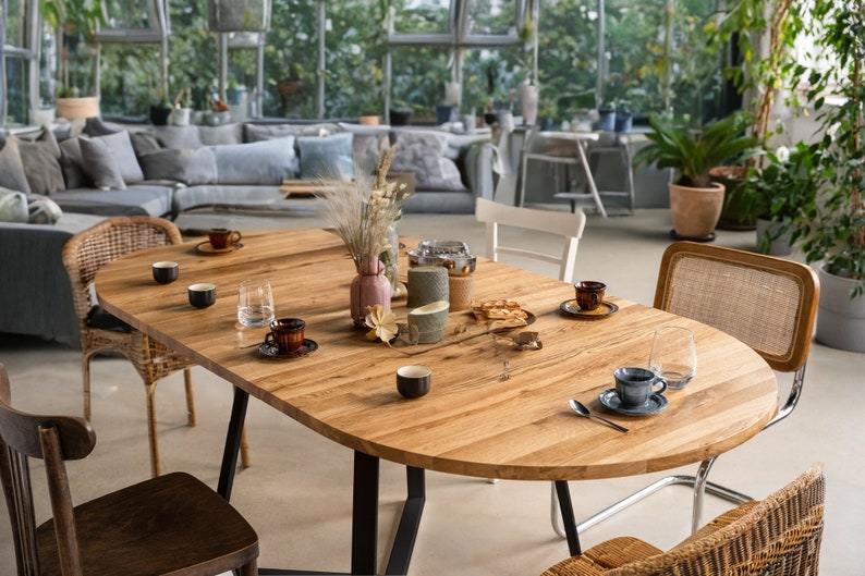 K&ouml;nnte beinhalten: Ein ovaler Holztisch mit schwarzen Metallbeinen, gedeckt mit vier St&uuml;hlen und einer Auswahl an Geschirr, darunter Tassen, Untertassen und ein Glas Wasser. Der Tisch ist von Gr&uuml;npflanzen und einer Couch im Hintergrund umgeben.
