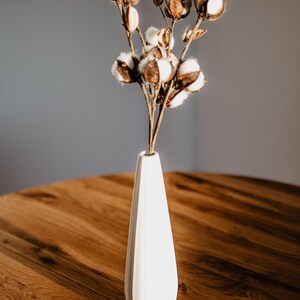 May include: A white ceramic vase with a fluted design holds a bouquet of dried cotton bolls. The vase is sitting on a wooden table.