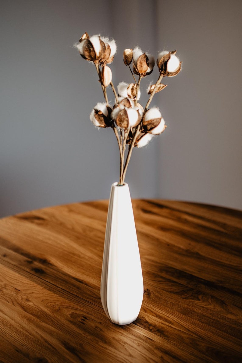 May include: A white ceramic vase with a fluted design holds a bouquet of dried cotton bolls. The vase is sitting on a wooden table.