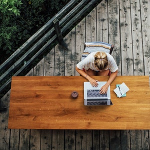 May include: Overhead view of a person working on a laptop at a wooden table on a deck. A cup and notebook are on the table. The deck is made of weathered wood, and lush greenery surrounds the deck.