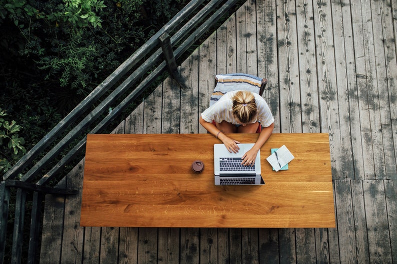 May include: Overhead view of a person working on a laptop at a wooden table on a deck. A cup and notebook are on the table. The deck is made of weathered wood, and lush greenery surrounds the deck.
