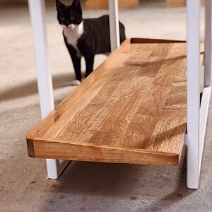 Puede incluir: Un marco de mesa de metal blanco con un estante de madera. El estante est&aacute; hecho de madera natural con un acabado marr&oacute;n claro. La mesa est&aacute; en una habitaci&oacute;n con un suelo blanco.