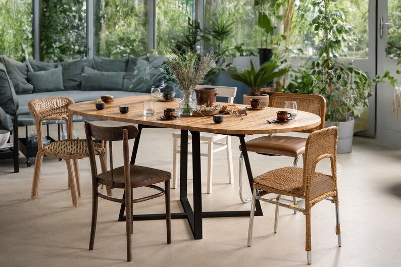 K&ouml;nnte beinhalten: Ein ovaler Esstisch aus Holz mit einem schwarzen Metallgestell, umgeben von sechs St&uuml;hlen. Der Tisch ist mit Geschirr, Tassen und einer Blumenvase gedeckt. Die St&uuml;hle sind in verschiedenen Stilen gehalten, darunter Korbgeflecht und Holz.